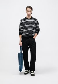 Black and gray striped sweater with a fuzzy texture, paired with black trousers and a blue tote bag. Model wears black shoes with white accents.