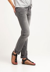 Jeans slim-fit grigi con orli arrotolati abbinati a sandali neri con cinturini, caratterizzati da una suola in sughero e dettagli in gomma.