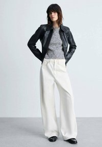 Giacca in pelle nera con colletto strutturato, maglione grigio a maglia, e pantaloni bianchi a gamba larga con cuciture visibili. Scarpe nere completano l'outfit.