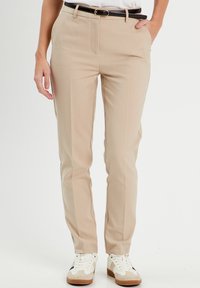 Beige, tailored trousers with a slim fit, featuring side pockets, a zip fly, and a black belt. Styled with white sneakers.