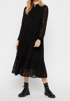 Vrouw draagt een zwarte midi-jurk met lange mouwen en kanten details, gecombineerd met zwarte veterlaarzen, tegen een effen witte achtergrond.