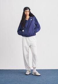 Tengerészkék könnyű dzseki fehér részletekkel, világosszürke melegítőnadrággal párosítva. A modell fekete sapkát és bézs sportcipőt visel logós részlettel.