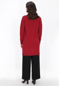 Pulover roșu cu mâneci lungi, cu slituri laterale și tiv cu nervuri, asortat cu pantaloni largi negri și tocuri negre. Textură netedă, croială lejeră.