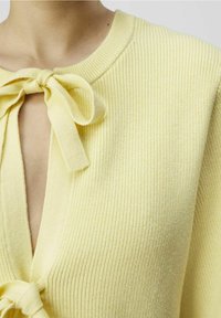 Top giallo in maglia con scollo a V e dettaglio con fiocco al collo. Il tessuto presenta una texture a coste e una finitura morbida.