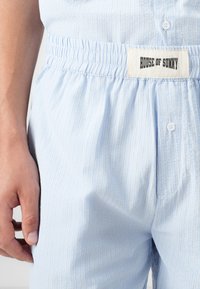 Hellblaue gestreifte Pyjamahose mit elastischem Bund und einem quadratischen Etikett mit der Aufschrift "HOUSE OF SUNNY". Weicher, atmungsaktiver Stoff mit Knopfdetails.