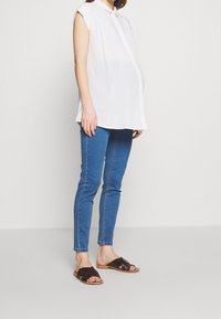 Top blanco sin mangas con cuello abotonado, combinado con jeans ajustados azules y sandalias marrones tipo slip-on.