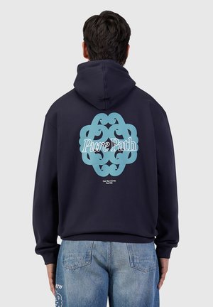 Persoon draagt een marineblauwe hoodie met een blauw abstract ontwerp en de tekst "Pure Path" op de rug, gecombineerd met lichtblauwe spijkerbroek.