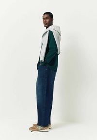 Szürke kapucnis pulcsi sötétzöld hosszú ujjú pólóra terítve, kék farmernadrággal és zöld kiegészítőkkel ellátott bézs tornacipővel párosítva.