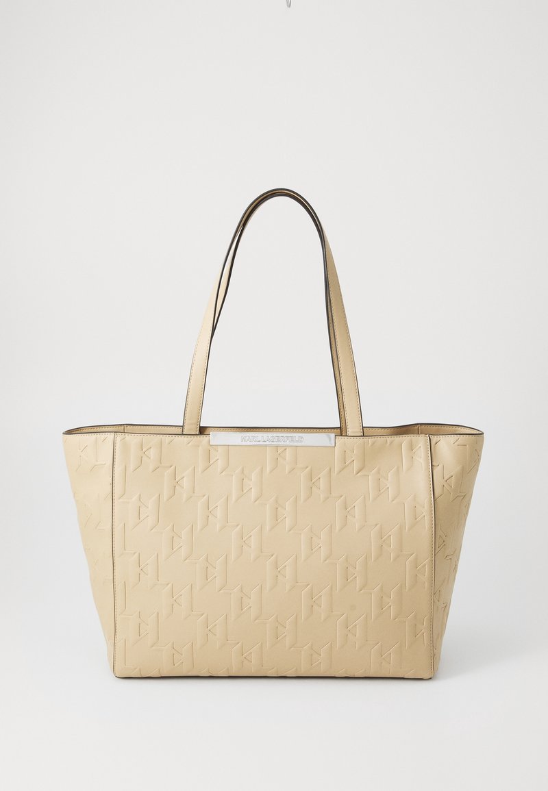 Sac cabas en simili cuir beige avec motif de logo embossé, poignées noires contrastantes, ouverture supérieure et plaque métallique de la marque à l'avant.