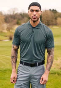 Polo shirt vert foncé avec des manches courtes et un col ; texture lisse. Assorti avec un pantalon gris clair et une ceinture noire, debout sur l'herbe.
