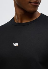 Juodas medvilninis marškinėlis su apvalia kaklo linija, ant kurio yra mažas baltas „BOSS“ logotipas ir švelnių spalvų. Lygus audinys, trumpomis rankovėmis.