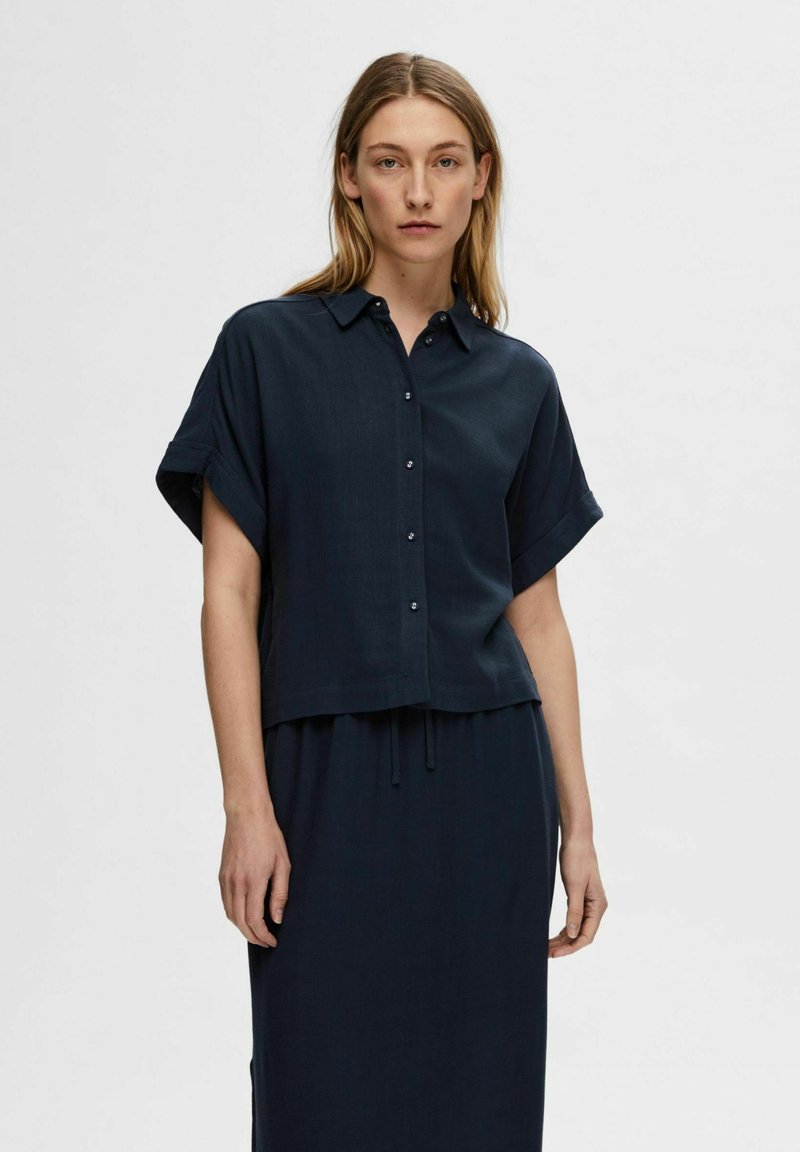 Camicia blu navy con maniche corte arrotolate, realizzata in tessuto leggero, presenta una vestibilità dritta e una sottile texture, abbinata a una gonna coordinata.