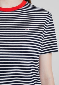 Camiseta de algodón con un patrón a rayas en azul marino y blanco, cuello rojo sólido y pequeño logotipo en el área del pecho izquierdo.