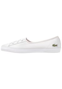 Baskets slip-on en toile blanche avec détails en lacets et logo de crocodile vert à l'arrière, semelle en caoutchouc plate, vue de côté sur fond blanc.