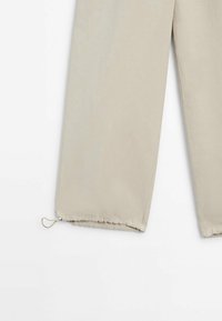 Beige-housut, joissa on sileä pinta, säädettävä nauha helmassa istuvuuden vuoksi ja suora lahkeensuukko.