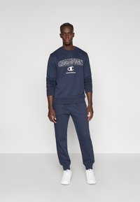 Μπλε φλις φούτερ με λογότυπο "CHAMPION", αντίστοιχο παντελόνι φόρμας και λευκά αθλητικά παπούτσια. Καθημερινά αθλητικά ρούχα με ριμπ μανσέτες και ελαστική μέση.