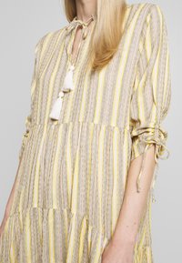 Robe rayée en beige, jaune et gris. Tissu léger avec un design à volants, encolure à nouer ajustable et accents de pompons au niveau du cou.