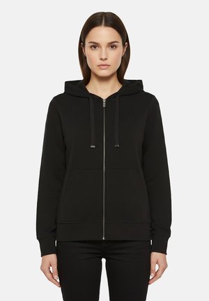 Donna che indossa una felpa con cappuccio nera con cerniera, coulisse e tasche frontali, in piedi contro uno sfondo chiaro e uniforme.