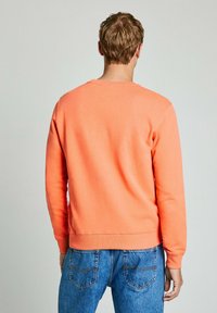Sudadera de algodón naranja con mangas largas, puños y dobladillo de canalé. Vista desde atrás, combinada con jeans azules.