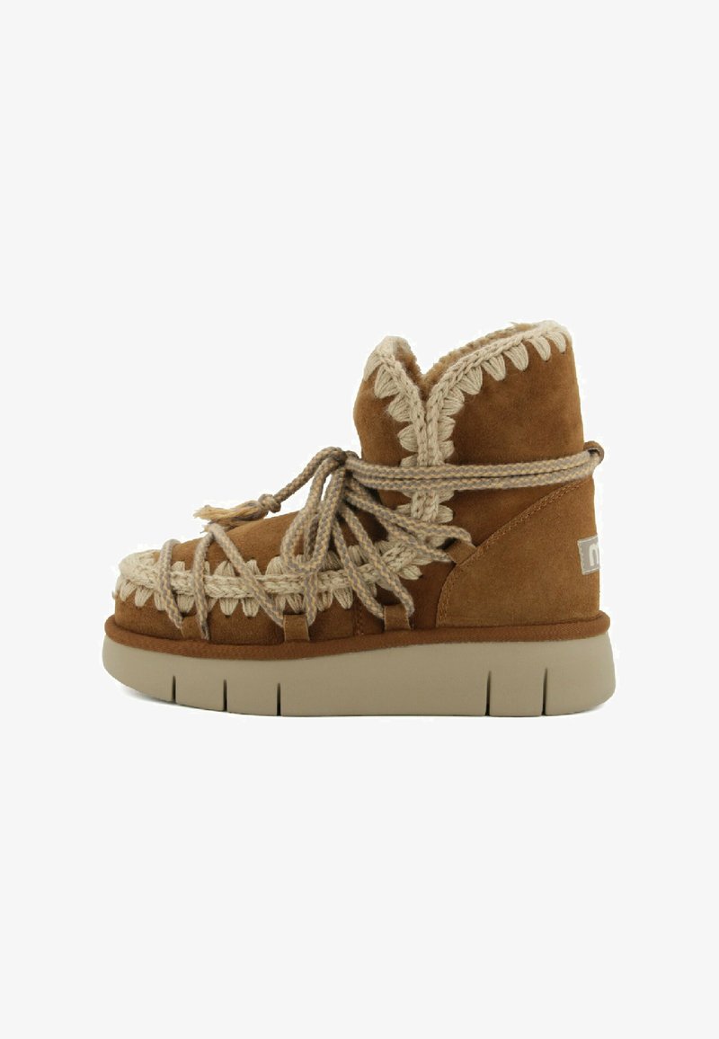 Des bottes de cheville en suede brun présentent un design texturé, des accents beiges tressés et une semelle épaisse couleur crème avec des rainures horizontales.