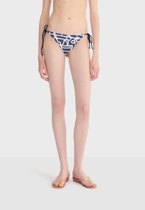 Woman wearing navy blue bikini bottoms with white abstract patterns and side ties, standing barefoot on a plain white background.