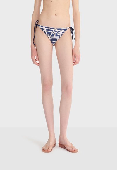 Femme portant un bas de bikini bleu marine avec des motifs abstraits blancs et des liens sur les côtés, debout pieds nus sur un fond blanc uni.