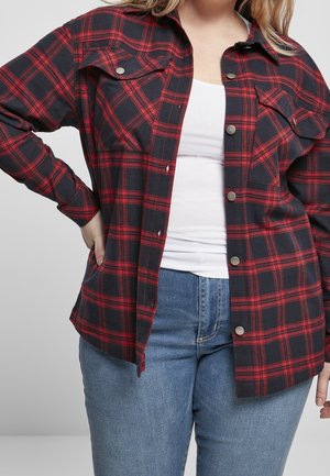 Frau trägt ein rot-schwarz kariertes Hemd über einem weißen Oberteil mit blauen Jeans, eine Hand in der Hüfte, vor einem einfarbigen Hintergrund.