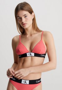 Bralette u koraljnom trokutu s tankim trakama i crnim elastičnim pojasom na kojem je istaknut logo "Calvin Klein". Glatka tekstura, minimalistički dizajn.
