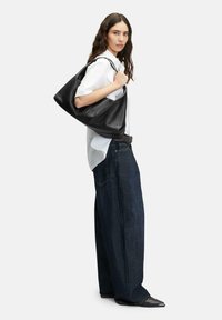 Czarna skórzana torebka hobo o luźnym fasonie, zakrzywionym kształcie, gładkiej fakturze i podwójnych ramiączkach; noszona z białą koszulą i szerokimi dżinsami.