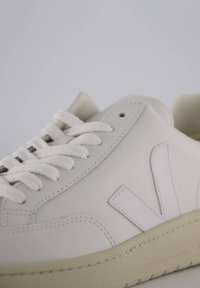 Hvide lædersneakers med en tekstureret overflade, der har en beige gummisål og en fremtrædende logoaccent på siden. Snørebåndene er hvide.