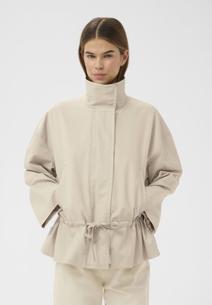 Žena v béžovej bunde s vysokým golierom, sťahovaním v páse a vreckami, stojaca pred jednoduchým svetlým pozadím.