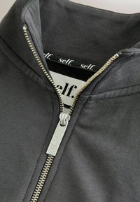 Szara kurtka na zamek z wysokim kołnierzem, srebrnym zamkiem błyskawicznym oraz czarną wewnętrzną metką z białym napisem "self.". Gładka bawełniana tkanina.