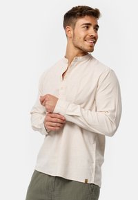 Camicia lunga beige chiara con colletto a camicia, dettagli in legno e vestibilità rilassata. Abbinata a pantaloni verdi testurizzati.