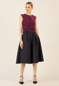 Spitzenoberteil in tiefem Lila mit gewellten Kanten, kombiniert mit einem marineblauen ausgestellten Rock. Besitzt eine taillierte Passform und schwarze spitze Absätze.