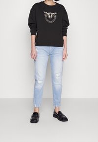 Person wearing black long-sleeve top with shiny bird design, light ripped jeans, and black loafers, standing against a plain background.