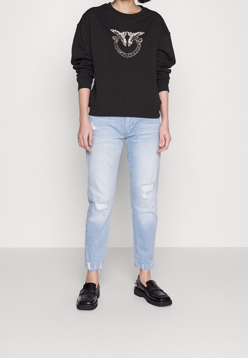 Person wearing black long-sleeve top with shiny bird design, light ripped jeans, and black loafers, standing against a plain background.