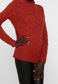 Stickad tröja i bränd orange, med ett upphöjt ribbat mönster, hög krage och långa ärmar med en texturerad yta.