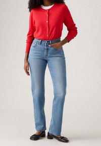 Cardigan rosso a maglia con chiusura a bottoni, indossato sopra una maglietta bianca. Jeans chiari a gamba diritta con vita alta, abbinati a ballerine nere.