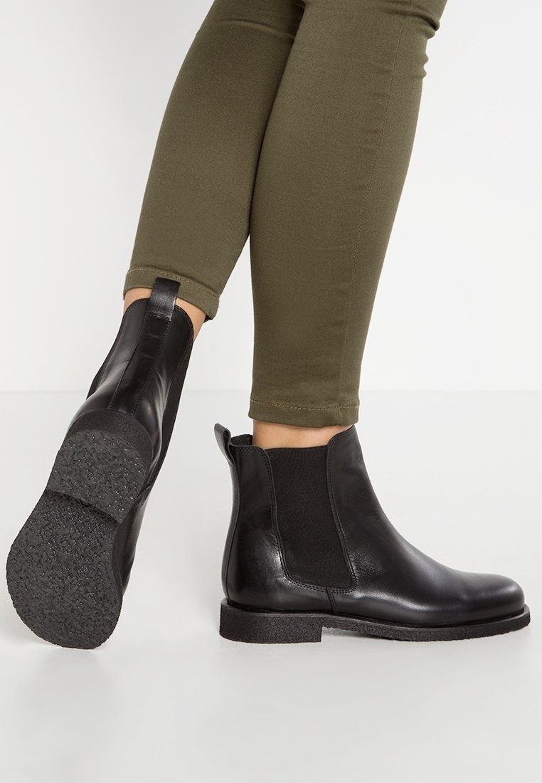 Personne portant des bottines Chelsea en cuir noir et un pantalon ajusté vert olive, debout sur un sol blanc, un pied levé montrant la texture de la semelle.