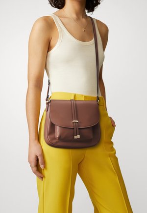 Sac bandoulière en cuir marron avec un rabat, deux sangles avant et des accessoires métalliques. Porté avec un débardeur crème et un pantalon jaune.