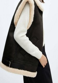 Veste sans manches en peau retournée marron avec un intérieur et un col de couleur crème, présentant une texture épaisse, une fermeture éclair et des poches latérales.