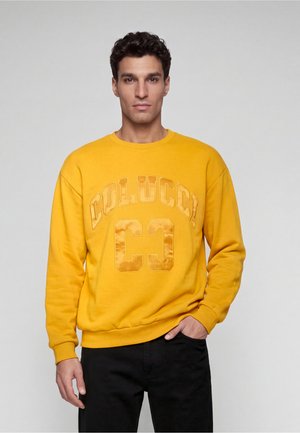 Uomo con felpa gialla con testo "COLUCCI CC" e pantaloni neri, in piedi davanti a uno sfondo grigio uniforme con una mano in tasca.