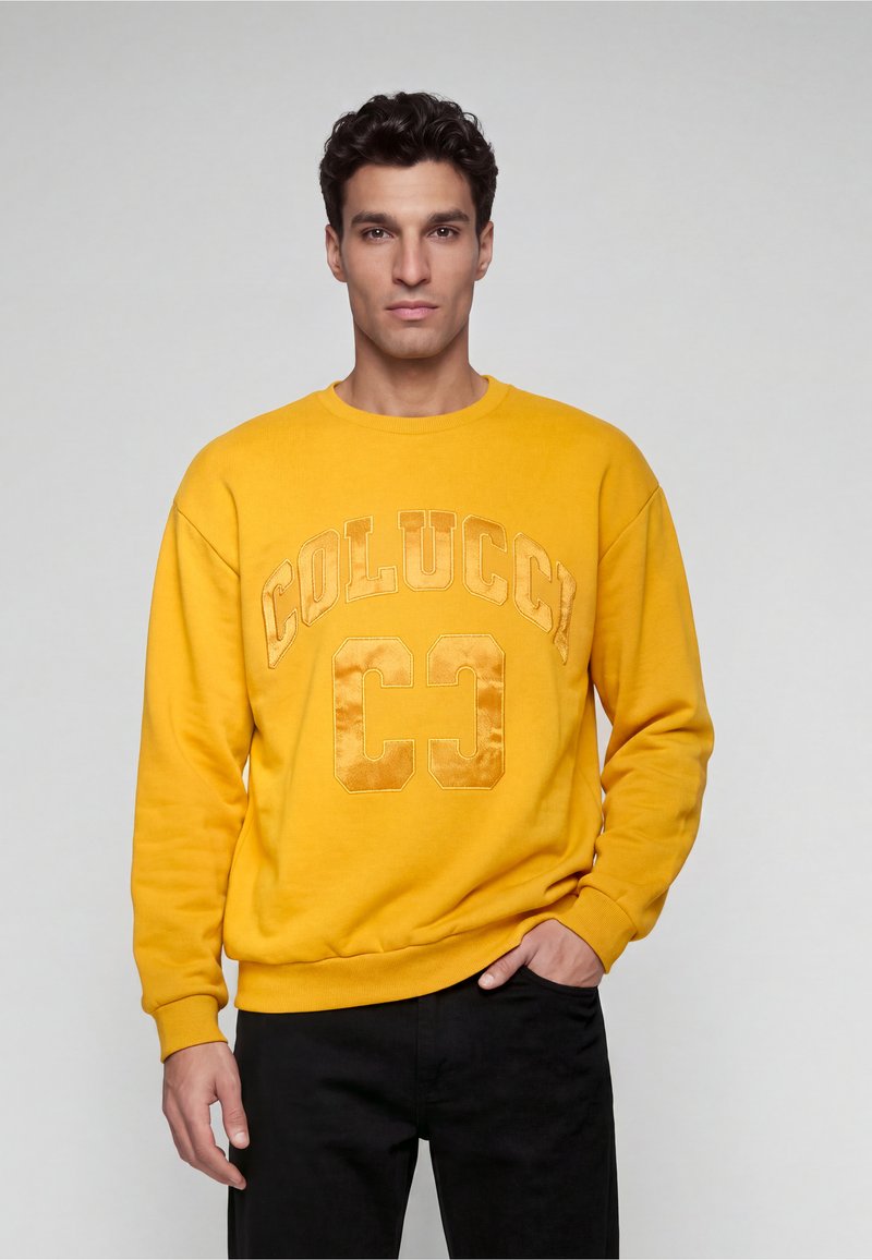 Homme portant un sweat jaune avec le texte "COLUCCI CC" et un pantalon noir, debout devant un fond gris uni avec une main dans la poche.
