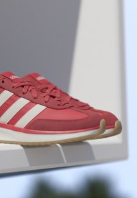 Baskets rouges Adidas avec bandes blanches et semelles en gomme disposées sur une étagère blanche sur un fond neutre.