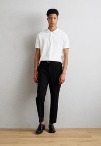Camisa polo blanca de manga corta, combinada con pantalones negros a medida y zapatos de cuero negros, sobre un fondo gris claro.