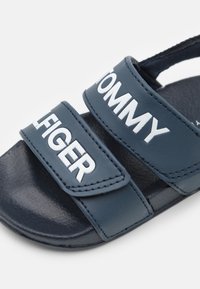 Navy modré sandály se dvěma nastavitelnými pásky s bílým textem 'TOMMY HILFIGER'. Texturovaná podrážka s hladkým koženým svrškem.