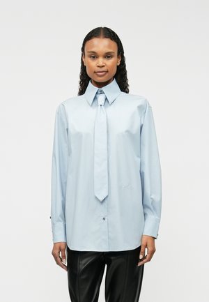 Cămașă albastră deschis cu guler ascuțit și cravată, având mâneci lungi și manșete cu nasturi, confecționată dintr-un material neted, asociată cu pantaloni negri.