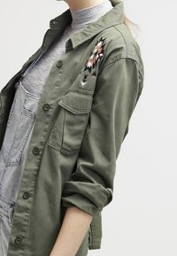 Chaqueta de camisa verde oliva con mangas largas, que presenta un bordado detallado en el hombro izquierdo en rosa, negro y blanco. Con frente abotonado y bolsillos.