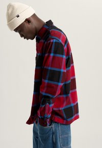 Rood, zwart en blauw geruit flanellen shirt met knoopmanchetten en een borstzak; gecombineerd met lichtblauwe denim jeans en een crèmekleurige beanie.