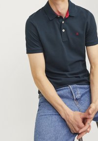 Námořnické polo tričko s krátkým rukávem, červeným vnitřním límcem a třemi knoflíky. Nosí se s světle modrými džíny, které zdůrazňují přiléhavý střih.